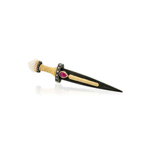 A miniature dagger or pin with a black blade, gold-toned guard and pommel, a white wrapped handle, and a small pink gemstone accent, shown on a clean white background with a faint reflection.