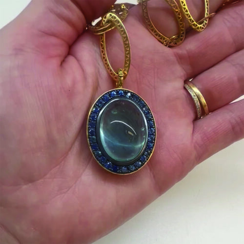 Large aquamarine cabochon pendant, 31.43 cts, with a pave-set blue sapphire surround in oxidized sterling silver, 2.70cttw set in 18k yellow gold crownwork with a hinged clasp.